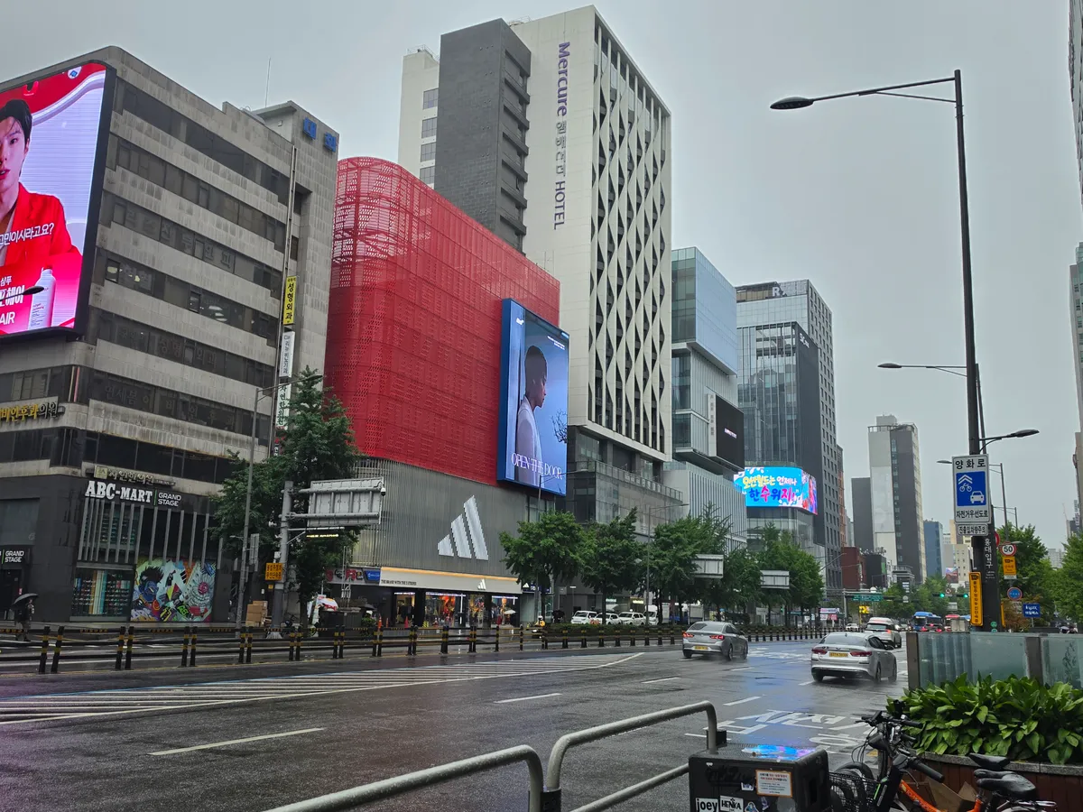 A rainy Hongdae streetscape along Yanghwa-ro, with storefronts, traffic and buildings.